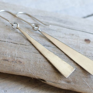 Può includere: Un paio di orecchini in argento a forma di triangolo con un design semplice. Gli orecchini sono appesi a una superficie in legno.