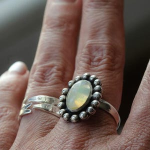 Peut inclure: Une bague en argent avec une pierre ovale de couleur claire, entourée de petites perles d'argent. Une autre bague en argent avec un motif de feuille est également visible. Les bagues sont sur le doigt d'une personne.