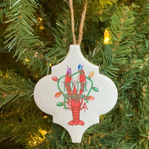 May include: White ceramic Christmas ornament shaped like a quatrefoil with a red lobster holding Christmas lights and the text "Tis the Season".