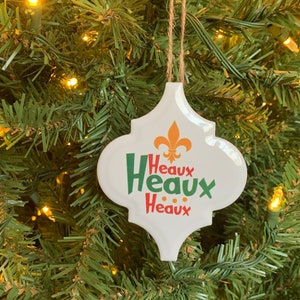 May include: White ceramic Christmas ornament with a green, red, and gold design. The ornament features the text "Heaux Heaux Heaux" and a fleur-de-lis.