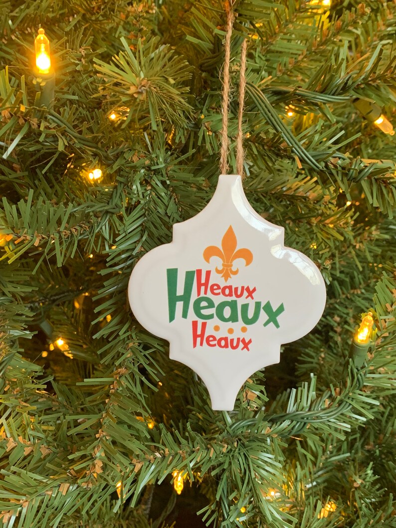 May include: White ceramic Christmas ornament with a green, red, and gold design. The ornament features the text "Heaux Heaux Heaux" and a fleur-de-lis.