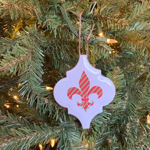 May include: A white ceramic Christmas ornament shaped like a fleur-de-lis with red and green striped accents.