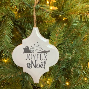 May include: White ceramic Christmas ornament with a black silhouette of a Santa Claus in a sleigh pulled by an alligator. The ornament reads "Joyeux Noel" in black lettering.