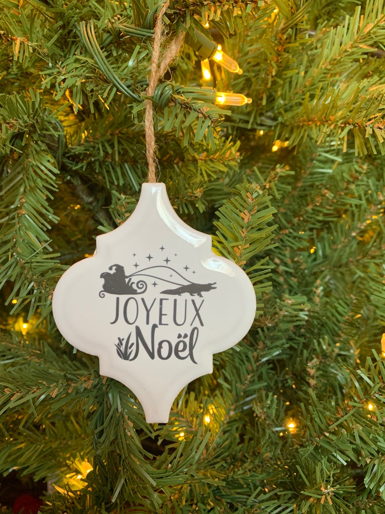 May include: White ceramic Christmas ornament with a black silhouette of a Santa Claus in a sleigh pulled by an alligator. The ornament reads "Joyeux Noel" in black lettering.