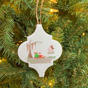May include: White ceramic Christmas ornament shaped like a quatrefoil with a painted scene of Santa Claus in a boat with a bag of gifts, a cypress tree with Spanish moss, and a small pink house.