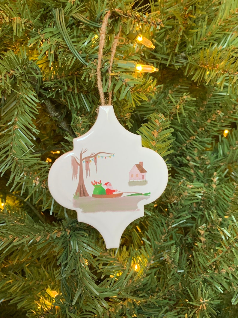 May include: White ceramic Christmas ornament shaped like a quatrefoil with a painted scene of Santa Claus in a boat with a bag of gifts, a cypress tree with Spanish moss, and a small pink house.