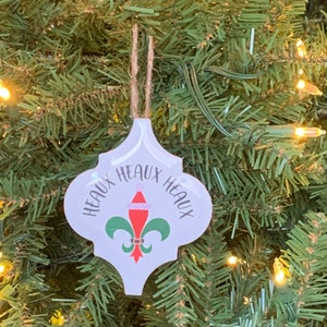 May include: White ceramic Christmas ornament with a green fleur-de-lis design and the text "Heaux Heaux Heaux" in black lettering. The ornament has a red Santa hat on top of the fleur-de-lis.