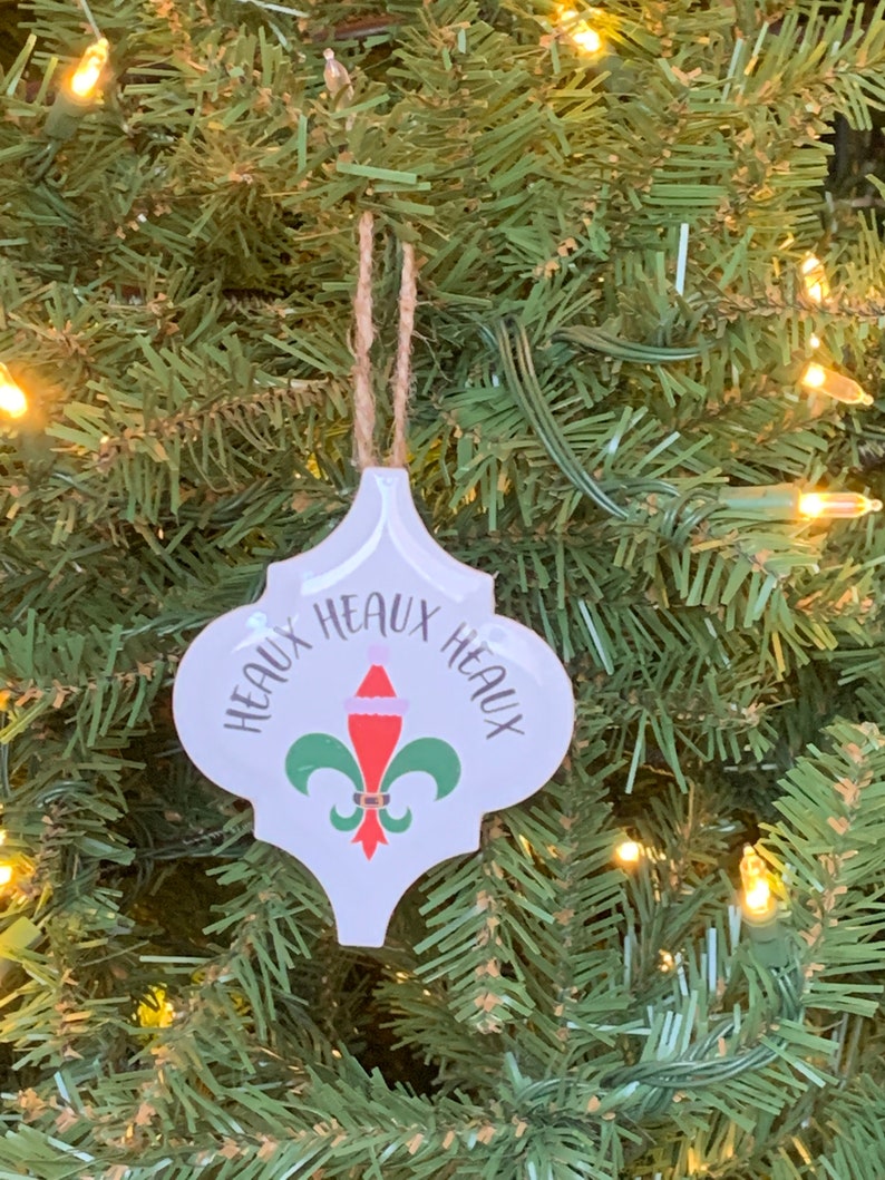 May include: White ceramic Christmas ornament with a green fleur-de-lis design and the text "Heaux Heaux Heaux" in black lettering. The ornament has a red Santa hat on top of the fleur-de-lis.