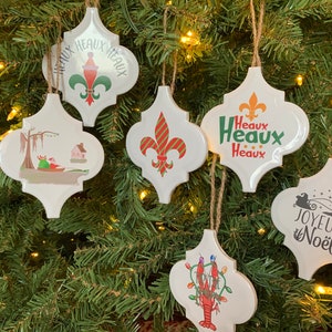 May include: Six white ceramic Christmas ornaments with different designs. The ornaments are shaped like a quatrefoil and have a string for hanging. The designs include a fleur-de-lis, a Christmas tree, a crawfish, a swamp scene, and the words "Joyeux Noel" and "Heaux Heaux Heaux".