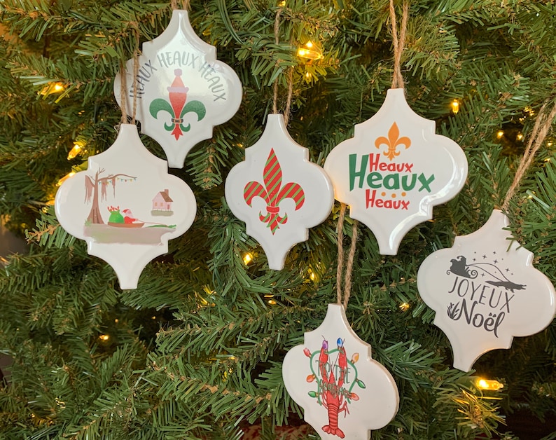 May include: Six white ceramic Christmas ornaments with different designs. The ornaments are shaped like a quatrefoil and have a string for hanging. The designs include a fleur-de-lis, a Christmas tree, a crawfish, a swamp scene, and the words "Joyeux Noel" and "Heaux Heaux Heaux".