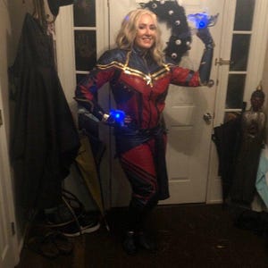 May include: A full-body Captain Marvel costume featuring a red, blue, and gold jumpsuit with long sleeves, a high collar, and a star emblem on the chest. The outfit includes matching gauntlets with glowing blue lights on the wrists and hands.