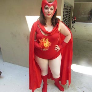 May include: A woman dressed as Scarlet Witch from the Marvel comics. She is wearing a red bodysuit with a red cape, a red headband with horns, and white boots. She is holding a small bouquet of orange and yellow flowers.