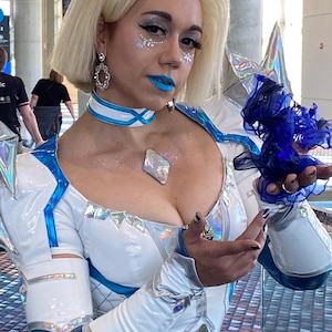May include: A white and metallic blue bodysuit featuring a high collar, iridescent diamond-shaped accents, and long sleeves. The outfit includes a matching choker, silver dangling earrings, and a dark blue sheer fabric accessory held in the hand.
