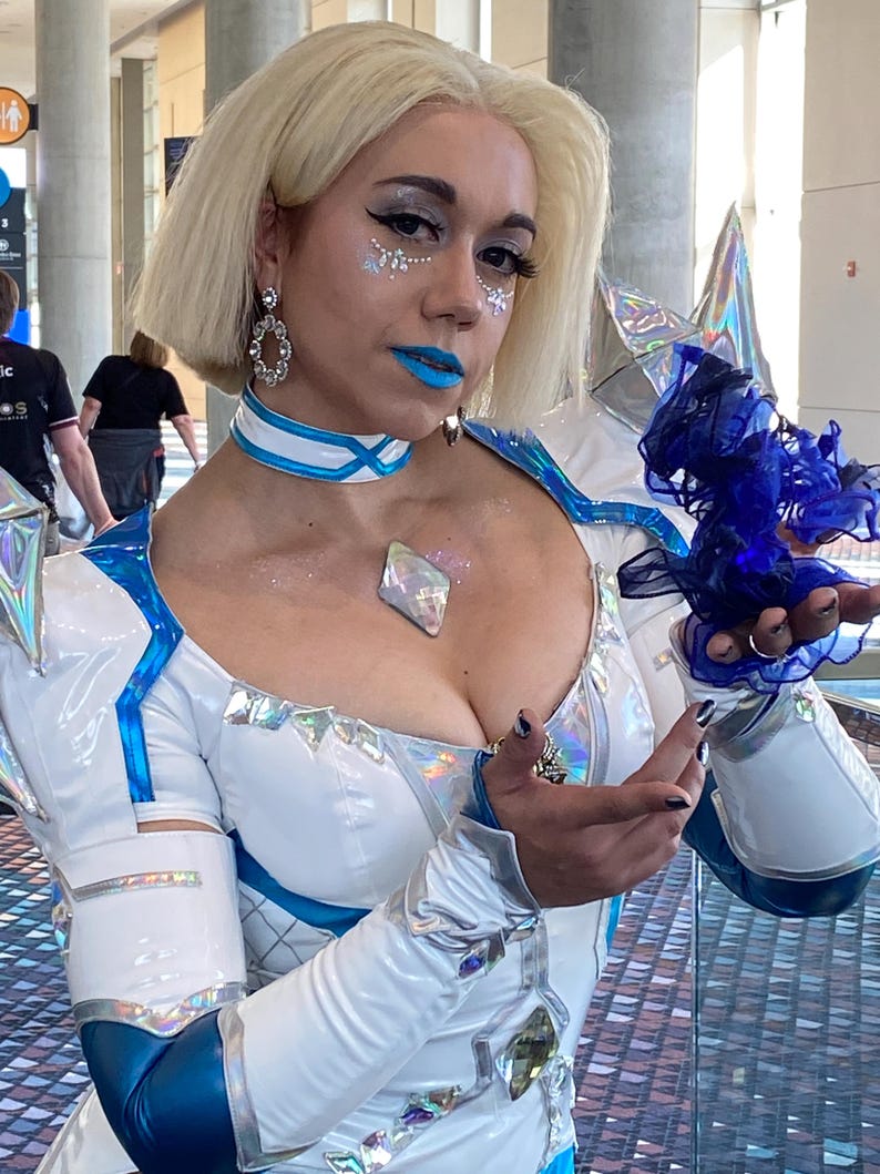May include: A white and metallic blue bodysuit featuring a high collar, iridescent diamond-shaped accents, and long sleeves. The outfit includes a matching choker, silver dangling earrings, and a dark blue sheer fabric accessory held in the hand.