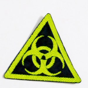 Könnte beinhalten: Ein gelber dreieckiger Patch mit einem schwarzen Biohazard-Symbol im Inneren. Das Symbol ist ein stilisiertes dreiblättriges Kleeblatt mit einem Kreis in der Mitte.