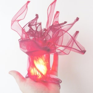 May include: A hand holding a small, glowing light wrapped in a pink tulle fabric. The fabric is gathered and ruffled, creating a whimsical and ethereal effect.