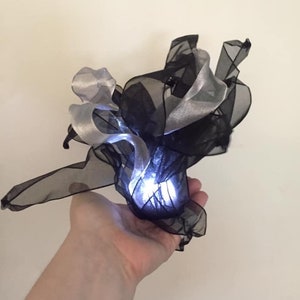 May include: A small, white light source is surrounded by black and silver sheer fabric. The fabric is gathered and ruffled, creating a cloud-like shape.