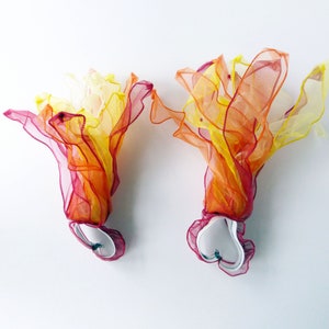 May include: Two pairs of colorful fabric dance streamers with white heart-shaped handles. The streamers are layered in shades of yellow, orange, and red.
