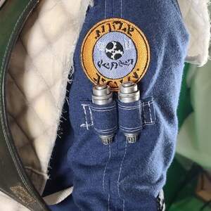 May include: A blue denim jacket with a white patch on the shoulder. The patch has a yellow circle with a blue circle inside. The blue circle has a gear and a symbol that looks like a cross. The text "Mandalorian" is written in a circular pattern around the blue circle.