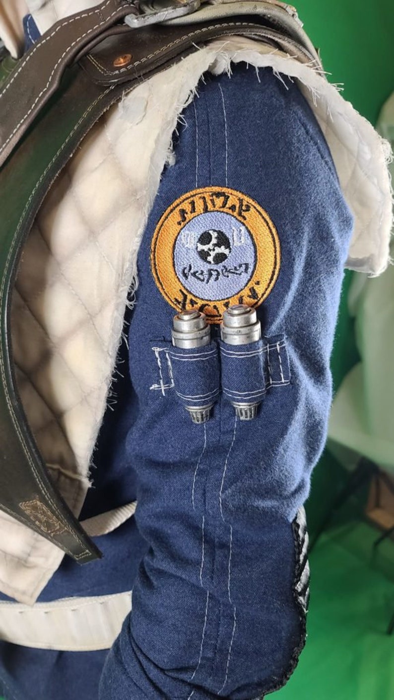 May include: A blue denim jacket with a white patch on the shoulder. The patch has a yellow circle with a blue circle inside. The blue circle has a gear and a symbol that looks like a cross. The text "Mandalorian" is written in a circular pattern around the blue circle.