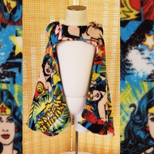 May include: A Wonder Woman fleece cape with a vibrant comic book print. The cape features images of Wonder Woman, stars, and comic book text on a blue, red, yellow, and beige background. The cape is displayed on a white mannequin.