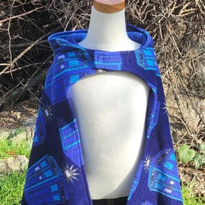 May include: A blue hooded cape with a pattern of blue and light blue rectangles and a white starburst design. The cape is made of a soft fabric and has a button closure at the neck. The cape is displayed on a white mannequin.