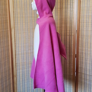 May include: A magenta shark costume cape with a hood, eye, and fin details. The hood features a white, toothy grin. The cape is displayed on a mannequin, showcasing its flowing design. The costume is made of a soft fabric.