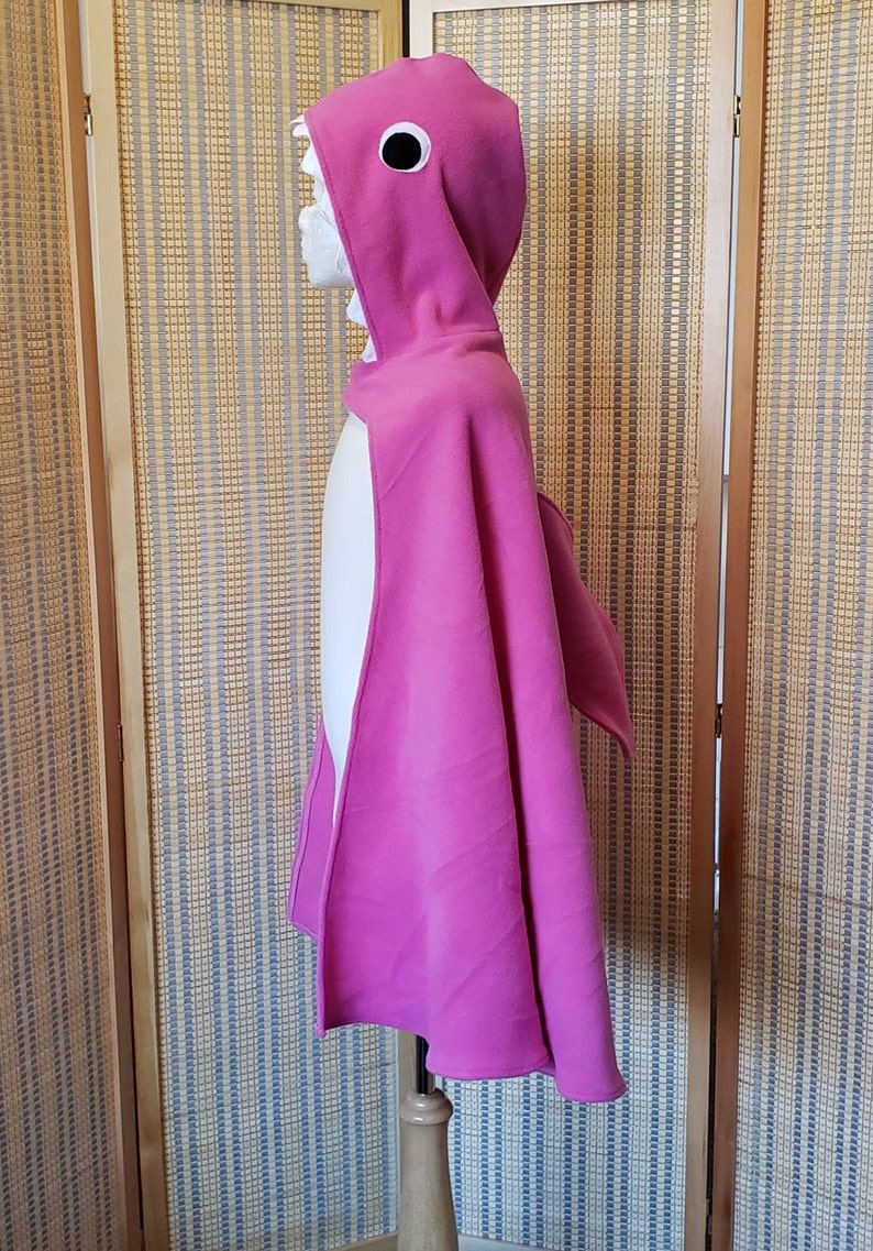 May include: A magenta shark costume cape with a hood, eye, and fin details. The hood features a white, toothy grin. The cape is displayed on a mannequin, showcasing its flowing design. The costume is made of a soft fabric.