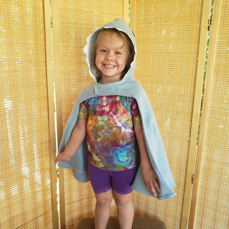 May include: A light blue hooded cape with a shark fin design, worn over a tie-dye t-shirt and purple shorts. The cape has a white lining and a snap closure. The child is smiling.