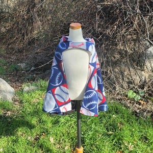 May include: A baseball-themed cape with a hood, displayed on a mannequin. The cape features a pattern of baseballs in red, white, and blue on a navy blue background. The cape is made of a soft, fleece-like material.