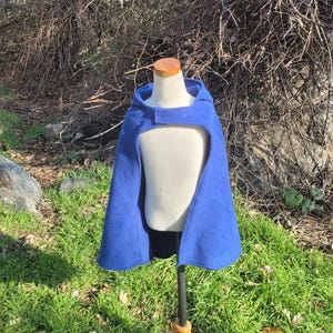 May include: A royal blue hooded cape displayed on a mannequin. The cape has a front opening with a velcro closure at the neck. The mannequin is on a wooden stand, set against a backdrop of green grass and natural foliage.