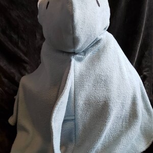 May include: A light blue hooded baby blanket. The blanket is made of a soft, plush material and features a hood with two small black eyes. The blanket is designed to resemble a bear or other animal. The blanket is shown on a dark brown surface.