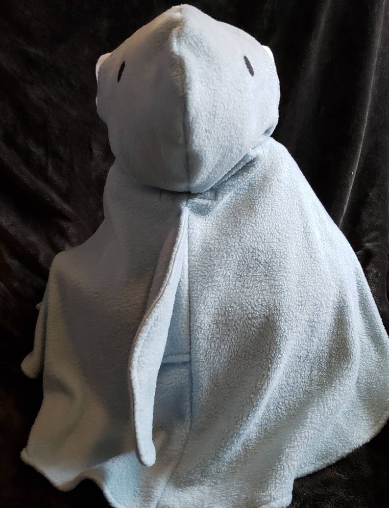 May include: A light blue hooded baby blanket. The blanket is made of a soft, plush material and features a hood with two small black eyes. The blanket is designed to resemble a bear or other animal. The blanket is shown on a dark brown surface.