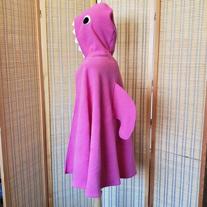 May include: A bright pink shark costume cape with a hood. The hood features a shark face with white teeth and a black eye. A large fin is attached to the side of the cape. The costume is displayed on a mannequin.