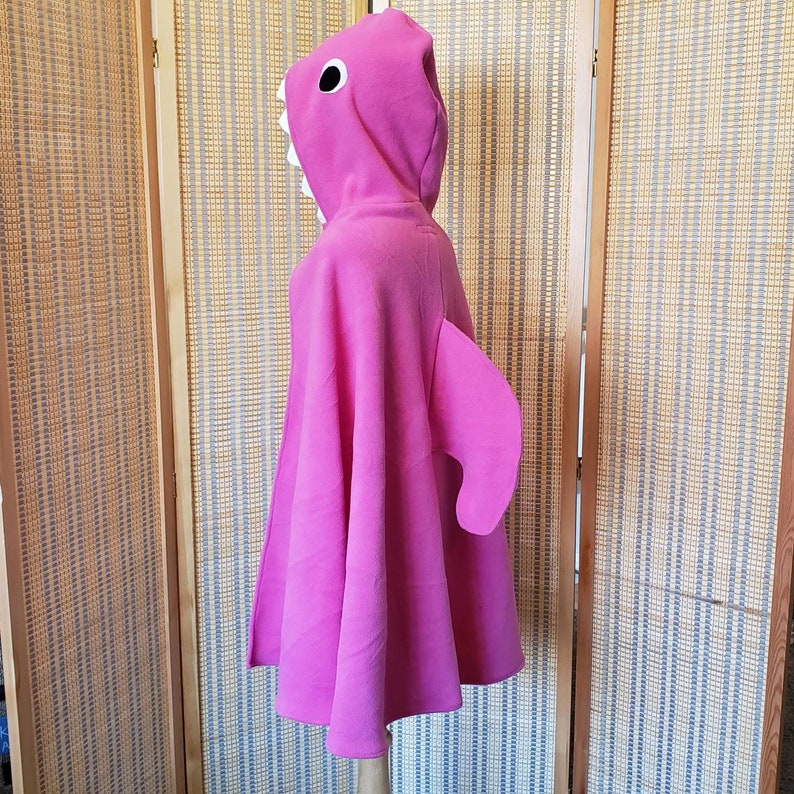 May include: A bright pink shark costume cape with a hood. The hood features a shark face with white teeth and a black eye. A large fin is attached to the side of the cape. The costume is displayed on a mannequin.