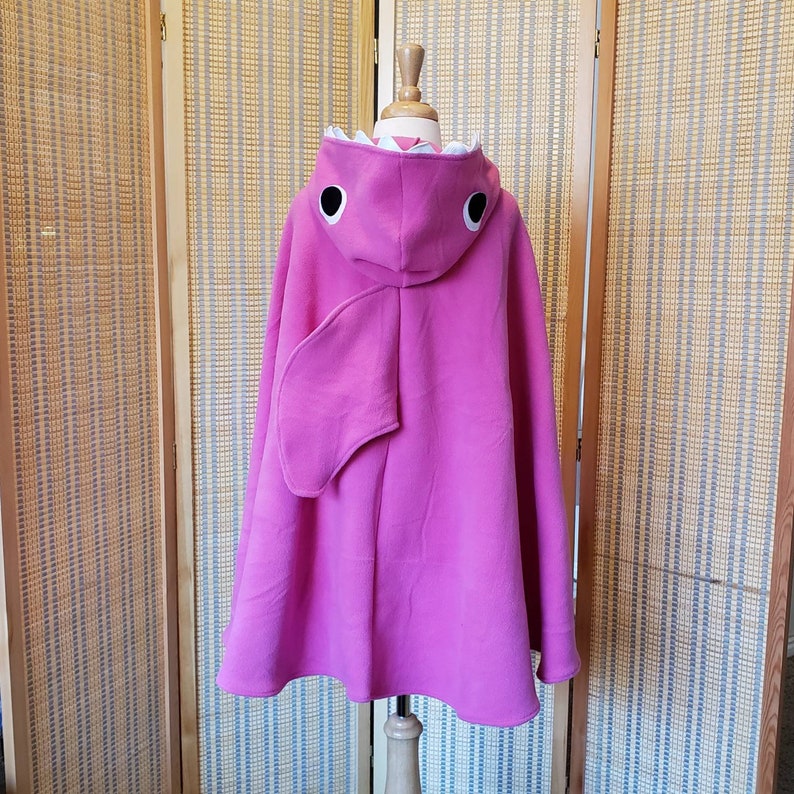 May include: A bright pink hooded cape, designed to resemble a shark. The hood features white teeth, black and white eyes, and a fin on each side. The cape is displayed on a mannequin.