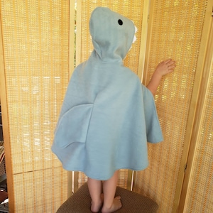 May include: A light blue shark-themed hooded cape. The cape features a hood with a shark face, including black eyes and white teeth. The cape has fin-shaped details on the sides. The child is standing on a brown woven stool.