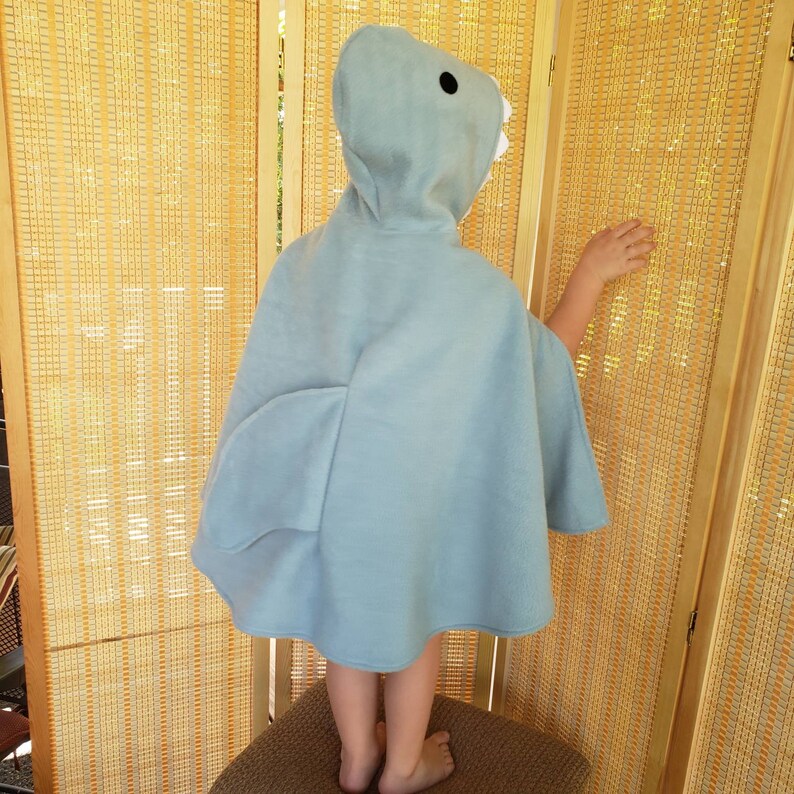 May include: A light blue shark-themed hooded cape. The cape features a hood with a shark face, including black eyes and white teeth. The cape has fin-shaped details on the sides. The child is standing on a brown woven stool.