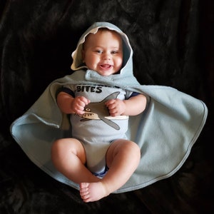May include: A baby wearing a light blue shark-themed hooded cape and a matching onesie. The onesie has a shark graphic and the text "Shark Bites". The baby is smiling and lying on a black surface.