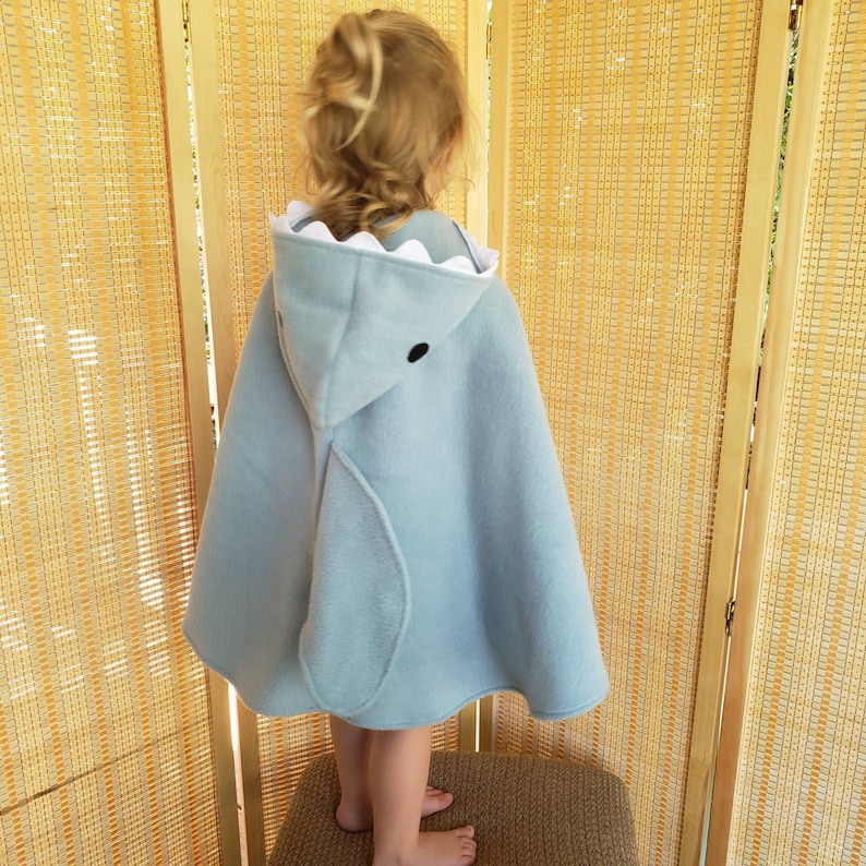 May include: A light blue shark-themed cape with a hood. The hood features a white, scalloped trim to resemble shark teeth and a black eye. The cape is made of soft material and is designed for children.