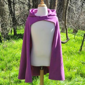 May include: A vibrant purple hooded cape with a button closure, displayed on a mannequin. The cape features a unique cutout design and is made of a soft, textured fabric. The background shows a natural outdoor setting with green grass and trees.