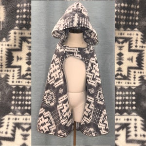 May include: A gray and white patterned hooded cape, displayed on a mannequin. The cape features a geometric design and is made of a soft, fleece-like material. The hood is up, and the cape is open in the front.