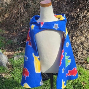 May include: A blue fleece cape with a hood, featuring colorful cartoon vehicle and star patterns. The cape has a yellow lining in the hood and a button closure at the neck. The cape is displayed on a mannequin.