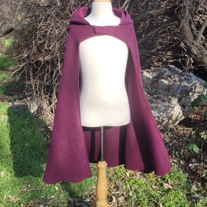 May include: A deep purple hooded cape displayed on a wooden mannequin. The cape has a button closure at the neck and a flowing design. The mannequin is set outdoors against a backdrop of trees and grass.