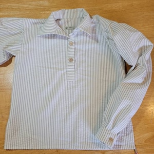 May include: A white and light grey striped shirt with a collar and long sleeves. The shirt has three buttons down the front and a button on each cuff. The text "little of" is visible on the collar. A pair of scissors is on the bottom right.
