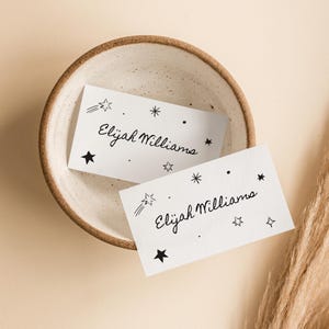 Puede incluir: Dos tarjetas blancas con tinta negra, cada una con el nombre "Elijah Williams" escrito en cursiva y una dispersión de pequeñas estrellas y una estrella fugaz.