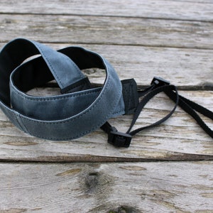 May include: A gray camera strap with black accents lies on a weathered wooden surface. The strap is made of a durable, canvas-like material and features adjustable black buckles and a loop for attachment.