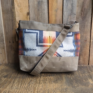 May include: A shoulder bag with a geometric pattern in shades of gray, orange, and blue. The bag has a solid olive-green base and trim, with a matching adjustable shoulder strap. The bag is made of a durable material and is suitable for everyday use.