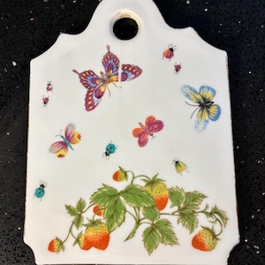 Può includere: Targa da parete in ceramica bianca con un design decorativo con farfalle colorate, coccinelle e fragole. La targa ha un foro nella parte superiore per appenderla. Le fragole sono rosse e gialle e le farfalle sono di vari colori.