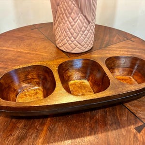 May include: A wooden serving dish with three oval compartments. The dish is made of dark brown wood and has a smooth, polished finish. A pink, textured vase is in the background. The dish is on a wooden table.